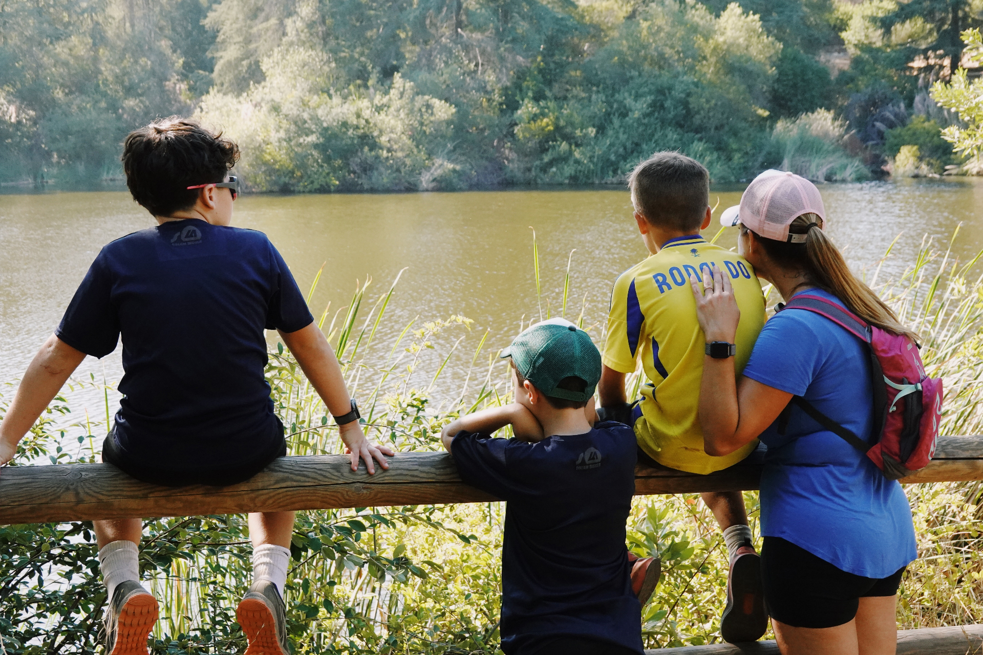 Three children and a guide sitting on a wooden fence looking out over a quiet lake